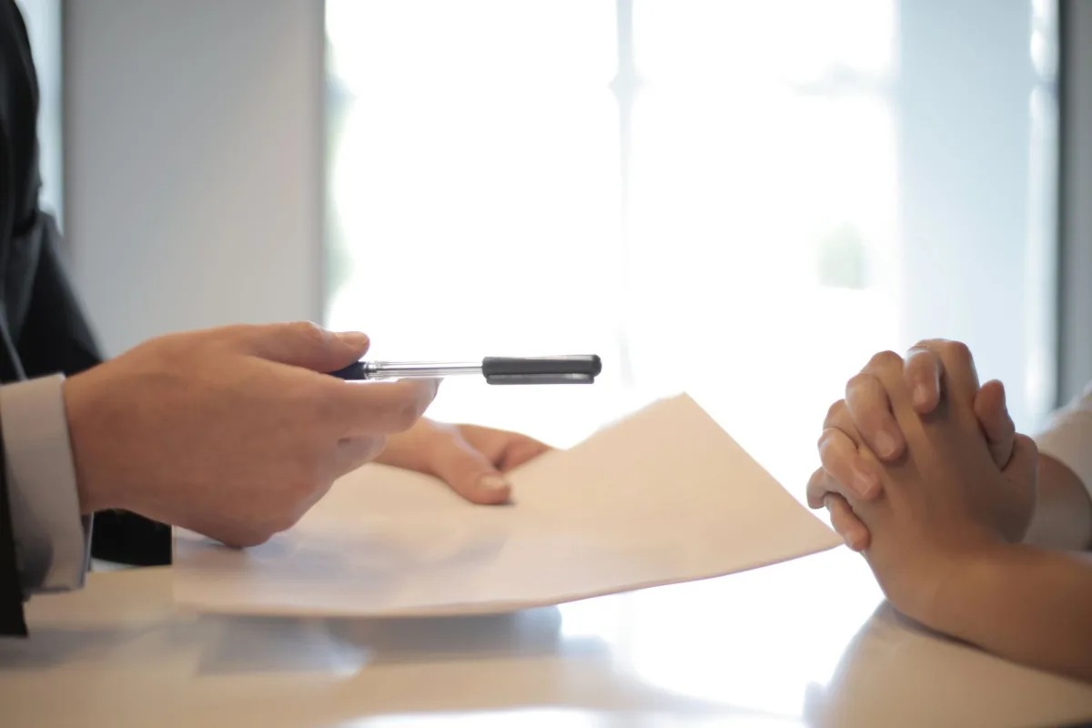 Side view of faceless formal man giving pen and paper to focused female with clenched hands at table on meeting Side view of faceless formal man giving pen and paper to focused female with clenched hands at table on meeting