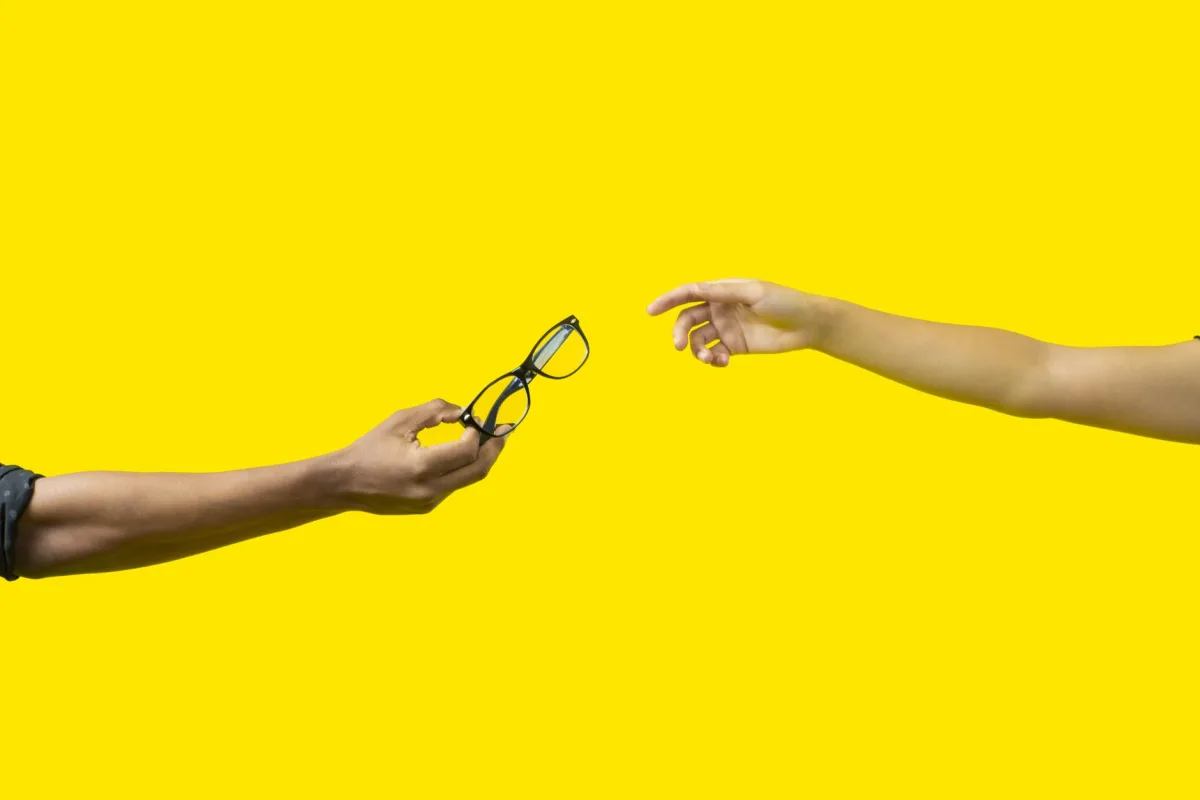 hands exchanging eyeglasses on a vibrant yellow backdrop, symbolizing connection.
