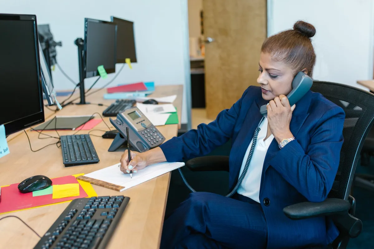 Businesswoman handling a phone while writing notes, surrounded by computers and office supplies.