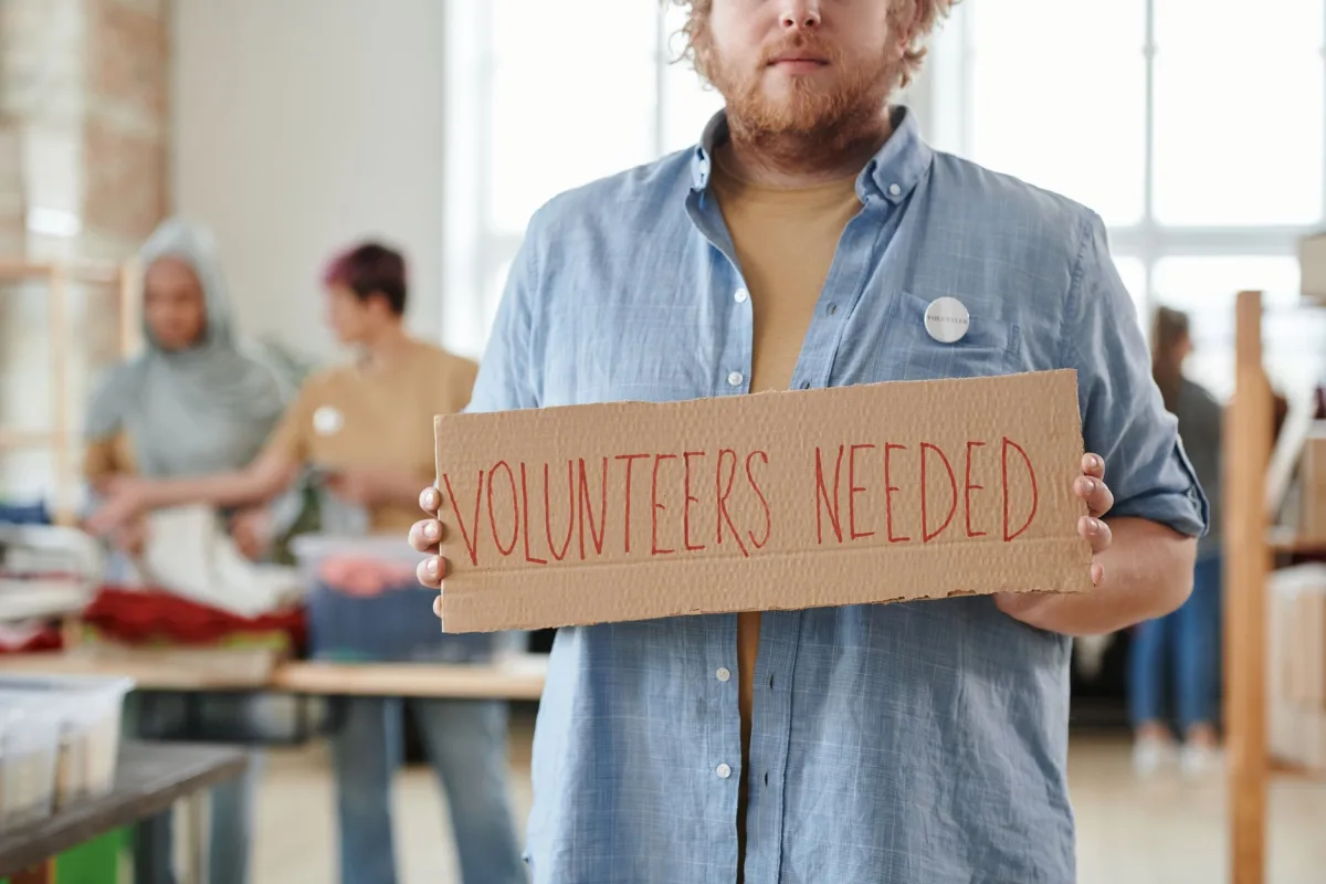 A man holds a 'Volunteers Needed' sign in a charitable organization setting, highlighting the for help.