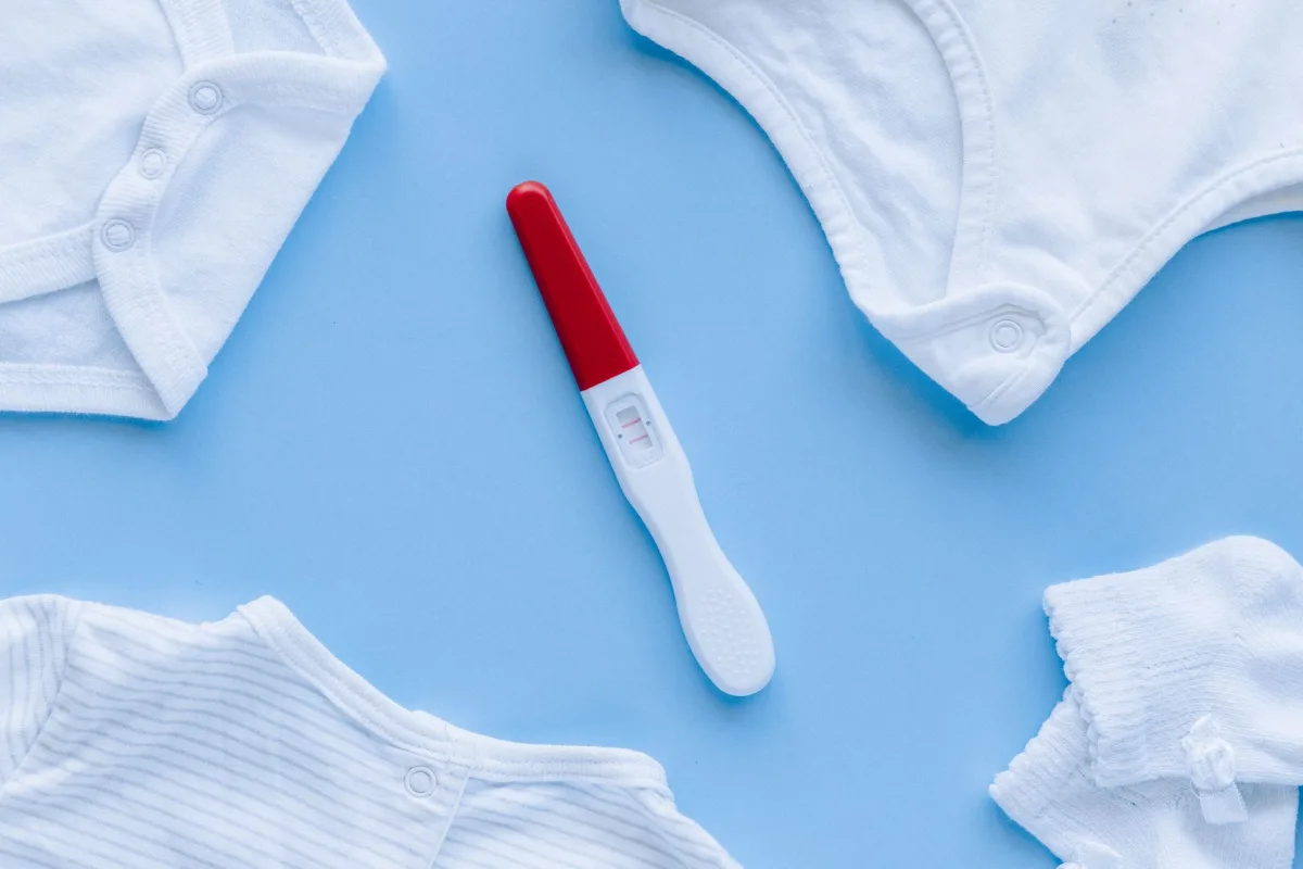 Flat lay of positive pregnancy test surrounded by baby clothes on blue background. Flat lay of positive pregnancy test surrounded by baby clothes on blue background.