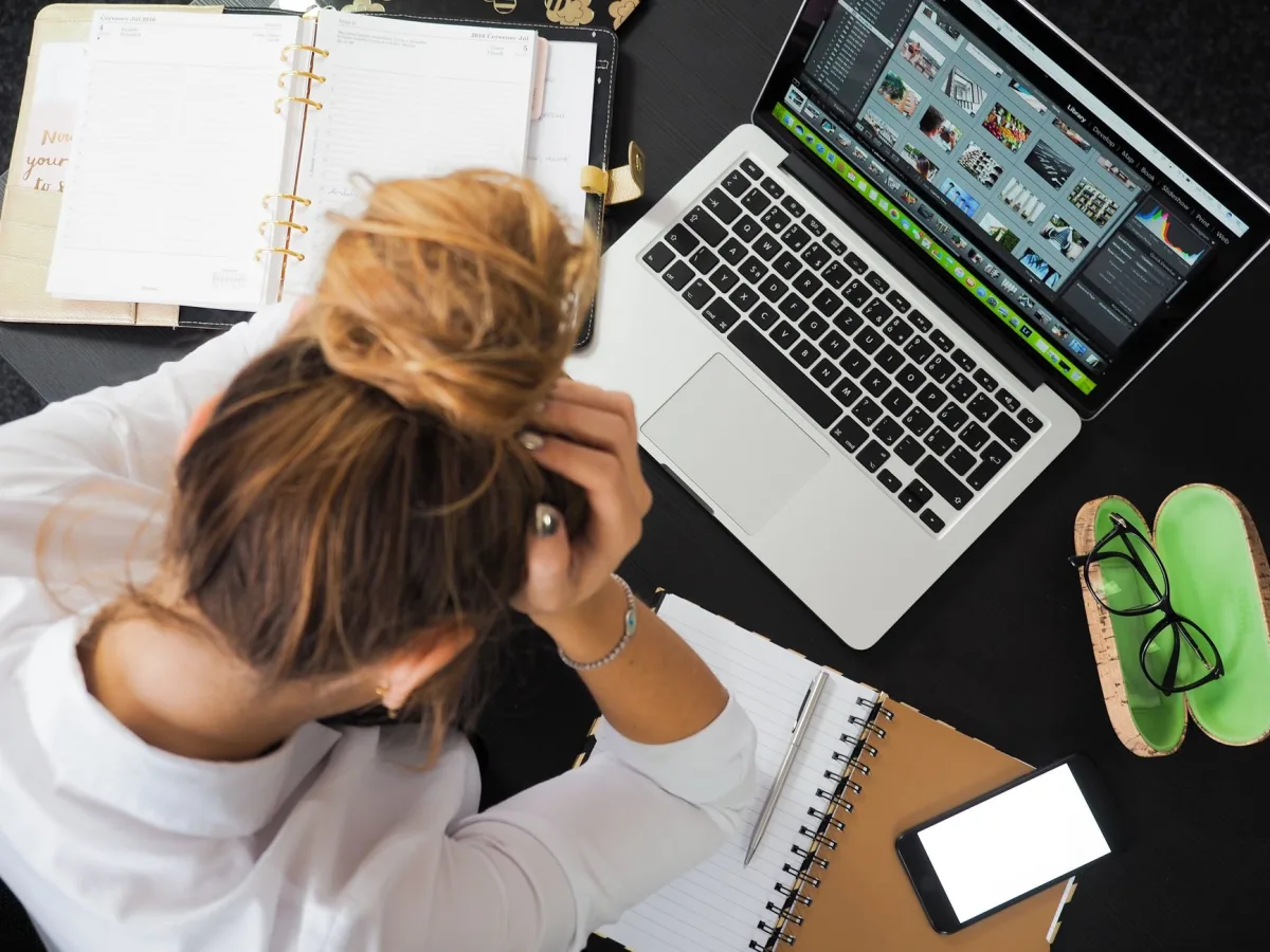 Overhead view of stressed woman working at desk with laptop, phone, and notebooks.